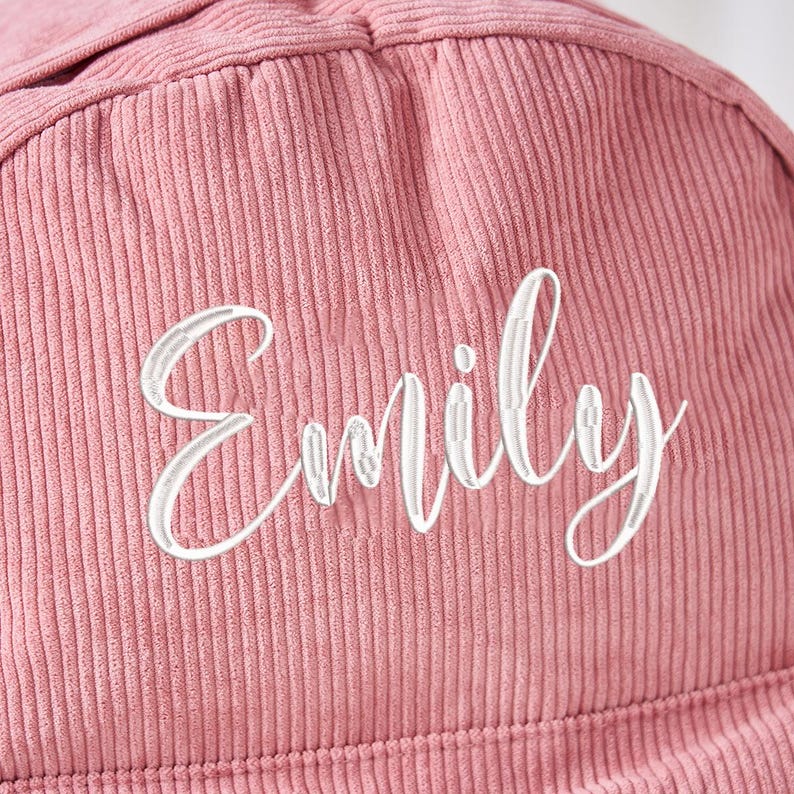 May include: Close-up of a dusty rose corduroy fabric with the name "Emily" embroidered in white cursive script. The fabric has vertical ridges, and the embroidery is centered.