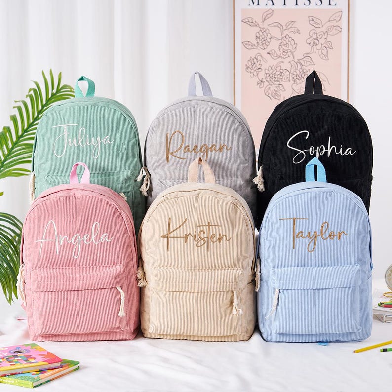 May include: A collection of corduroy backpacks in various colors, including pink, mint green, gray, black, and light blue. Each backpack features a front pocket, top handle, and the name of a person in cursive script. The backpacks are suitable for school or travel.