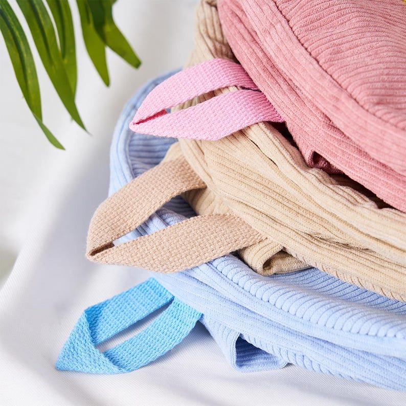 May include: Close-up of several corduroy tote bags in pastel colors: pink, beige, and light blue. Each bag has a matching fabric handle. The bags are stacked on top of each other, with the handles visible.