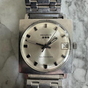May include: A vintage Benrus selfwinding wristwatch with a square silver-tone case and a matching metal band. The watch face is silver with black markers, hands, and the date displayed at the 3 o'clock position. The watch has the text "BENRUS" and "SELF WINDING".