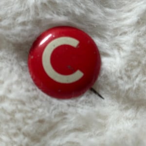 May include: A red and white enamel pin with the letter "C" in white on a red background.