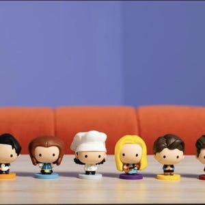May include: Six cartoon figurines of characters from the TV show Friends. Each figure has a unique hairstyle and outfit, standing on a coloured base. The background features a blue wall and an orange sofa.