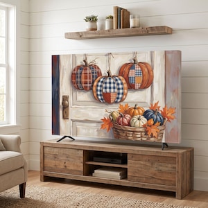 May include: A television screen displaying a painting of three hanging pumpkins with plaid and checkered patterns above a woven basket filled with smaller pumpkins and orange autumn leaves, all set against a white paneled door background.