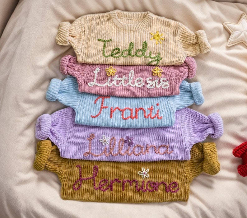 May include: A stack of five knitted jumpers with embroidered names, "Teddy", "Little Sis", "Franti", "Liliana", and "Hermione". Each jumper has a different colour and features embroidered flowers.