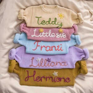 May include: A stack of five knitted sweaters with embroidered names, including "Teddy", "Little Sis", "Franti", "Lilliana", and "Hermione". Each sweater has a different color and features small embroidered flowers.