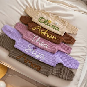May include: A stack of five knitted sweaters in various colors, each with a different name embroidered on the front. The names are Olivia, Asher, Thea, Siya, and Amess.