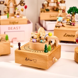 May include: Wooden music box featuring two bear figurines on a circular platform with a train track design. The box has a crank and is decorated with miniature trees and a small wooden structure. The name "Asher" is inscribed on the front.