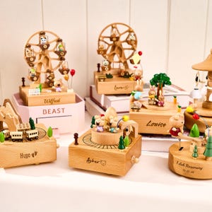 Op de afbeelding: Een verzameling houten muziekdozen met verschillende thema's. De dozen hebben reuzenraderen, treinen en carrousels, elk versierd met miniatuur dierenfiguren en decoratieve elementen. Sommige dozen hebben namen gegraveerd.