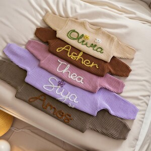 May include: A stack of five knitted jumpers in various colours, each with a different name embroidered on the front. The names are Olivia, Asher, Thea, Siya, and Amess.