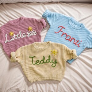 May include: Three knitted jumpers with embroidered names. A pink jumper with "Little sis" embroidered in white, a light blue jumper with "Franti" embroidered in red, and a cream jumper with "Teddy" embroidered in green.