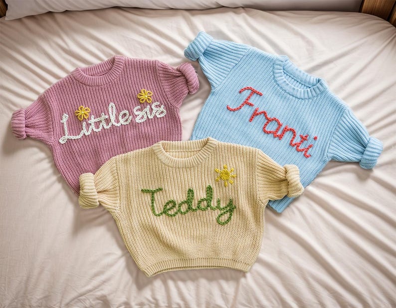 May include: Three knitted jumpers with embroidered names. A pink jumper with "Little sis" embroidered in white, a light blue jumper with "Franti" embroidered in red, and a cream jumper with "Teddy" embroidered in green.