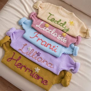 May include: A stack of five knitted jumpers in various pastel colours, each with a different name embroidered on the front. The names are "Hermione", "Liliana", "Franti", "Little Sis", and "Teddy".