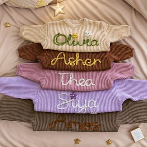 May include: A stack of six knitted jumpers in various colours, each with a different name embroidered on the front. The names are Olivia, Asher, Thea, Siya, and Ames.