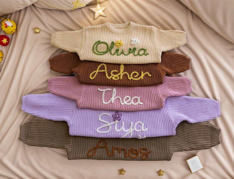 May include: A stack of six knitted jumpers in various colours, each with a different name embroidered on the front. The names are Olivia, Asher, Thea, Siya, and Ames.