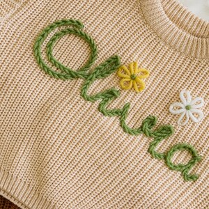 May include: A beige knitted jumper with the name "Olivia" embroidered in green yarn and decorated with yellow and white flowers.