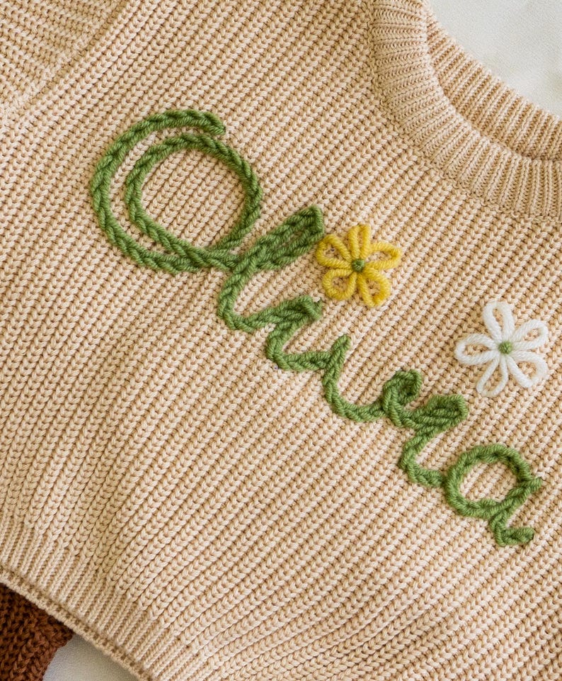 May include: A beige knitted jumper with the name "Olivia" embroidered in green yarn and decorated with yellow and white flowers.