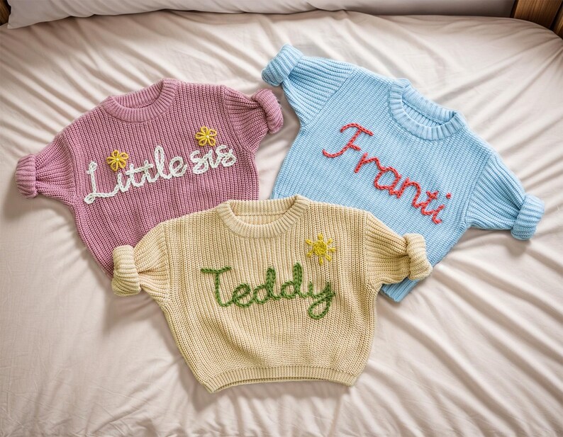 May include: Three knitted sweaters with embroidered names. The sweaters are pink, light blue, and cream. The names are "Little sis", "Franti", and "Teddy".