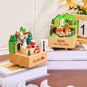 May include: Two wooden Christmas-themed music boxes. One features a Santa Claus figure, a snowman, and a train. The other has a train, houses, and a windmill. Both have personalized text. The boxes are on top of books.