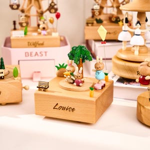 Pu&ograve; includere: Carillon in legno con un tema giocoso. La scatola centrale presenta una famiglia di conigli intorno a un albero, con il nome "Louise" inciso. Altre scatole includono una ruota panoramica, una giostra e una piccola panchina.