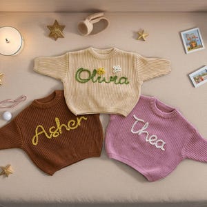 May include: Three knitted baby sweaters with embroidered names: Olivia, Asher, and Thea. The sweaters are in cream, brown, and pink.