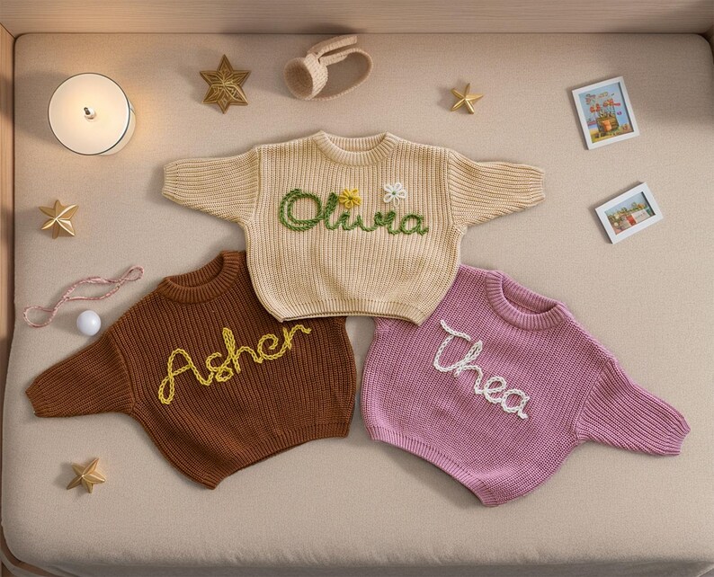 May include: Three knitted baby sweaters with embroidered names: Olivia, Asher, and Thea. The sweaters are in cream, brown, and pink.