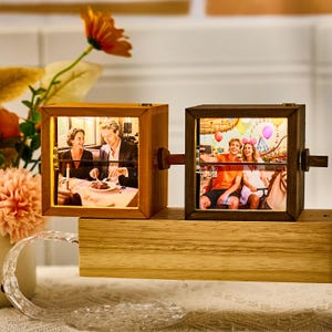 May include: Two wooden photo cubes linked by a wooden arm. Each cube displays a backlit photograph. One shows a couple at a restaurant, the other shows a couple on a carousel. The cubes sit on a wooden base.