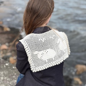Puede incluir: Un cuello de ganchillo rectangular de color crema con un patrón de rejilla y dos motivos de ovejas integrados en el diseño. El accesorio se lleva sobre una americana gris carbón oscuro y presenta un borde festoneado en la parte inferior.