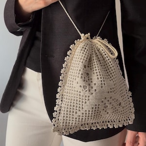 Puede incluir: Una bolsa con cordón de color crema confeccionada en encaje de ganchillo intrincado con un patrón de cuadrícula cuadrada y un borde festoneado decorativo. El pequeño bolso cuenta con un cierre de cordón a juego y una fina correa de ganchillo.