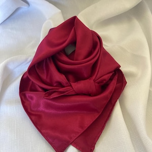 Puede incluir: Una bufanda de satén rojo oscuro anudada. La bufanda está doblada en forma triangular y está sobre una superficie de tela blanca.