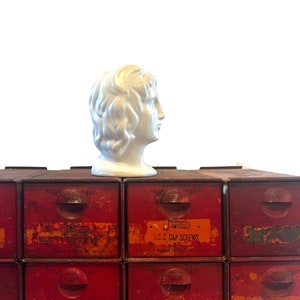 May include: A white ceramic bust of a woman with a long, flowing hairstyle sits on top of a set of red metal drawers. The drawers have labels that read "S.A.E. CAP SCREWS" and "U.S.S. CAP SCREWS".