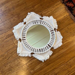 May include: A white macrame mirror with a round mirror in the center. The mirror is surrounded by a white macrame design with fringe.