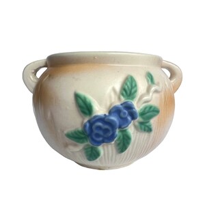 Pode incluir: Um vaso de cerâmica branco com detalhes florais azuis e verdes. O vaso tem duas alças e uma forma arredondada.