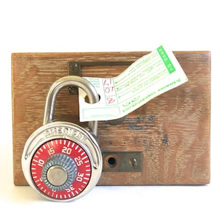Pode incluir: Um cadeado de combinação prateado com um mostrador vermelho e a palavra "American" impressa nele. O cadeado está preso a uma caixa de madeira com uma etiqueta que diz "American Combination Lock".