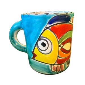 May include: A brightly coloured ceramic mug with a fish design. The mug has a blue handle and a fish illustration in yellow, red, green, and orange. The mug's exterior is primarily blue and teal, with a textured rim. The mug is suitable for hot or cold drinks.