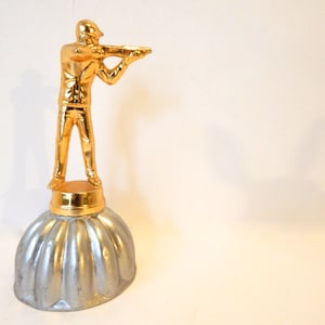 Puede incluir: Un trofeo dorado con una figura de una persona sosteniendo un rifle, de pie sobre una base plateada con un diseño acanalado.