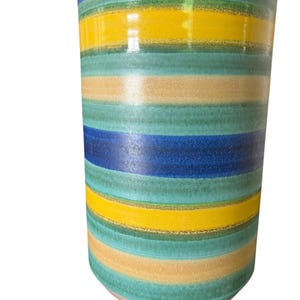 May include: A cylindrical ceramic vase with horizontal stripes in shades of blue, green, yellow, and tan. The vase has a glossy finish and a light-colored base. The design is reminiscent of a retro or mid-century modern style.