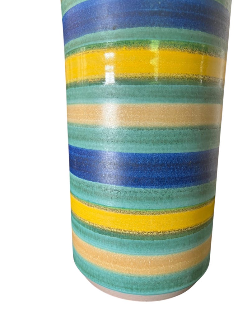 May include: A cylindrical ceramic vase with horizontal stripes in shades of blue, green, yellow, and tan. The vase has a glossy finish and a light-colored base. The design is reminiscent of a retro or mid-century modern style.