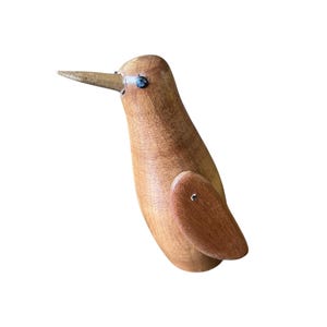 May include: A wooden bird ornament with a long beak and small, dark eyes. The bird is made of light brown wood and has a simple, minimalist design. The wings are attached with small metal pins. A decorative item.