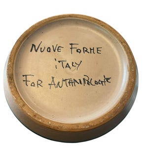 May include: A ceramic bowl's interior, featuring handwritten text in black ink. The text reads "NUOVE FORME ITALY For ANTHROPOLOGIE". The bowl has a light brown interior and a darker brown rim.