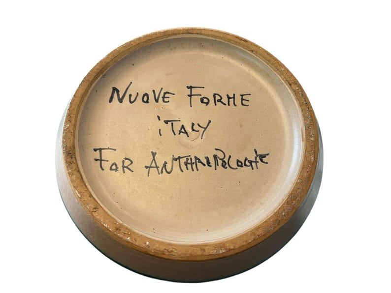 May include: A ceramic bowl's interior, featuring handwritten text in black ink. The text reads "NUOVE FORME ITALY For ANTHROPOLOGIE". The bowl has a light brown interior and a darker brown rim.