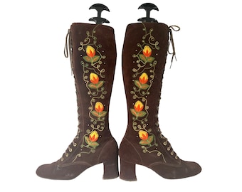 May include: Brown suede knee-high boots with a floral embroidery design featuring yellow, orange, and green flowers. The boots have a lace-up front and a block heel.