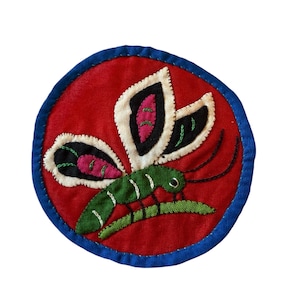 May include: A round, embroidered patch featuring a stylized insect. The insect has a green body, white and black wings with pink accents, and is set against a red background with a blue border. The patch is likely for decorative purposes.