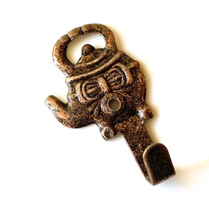 Puede incluir: Un gancho de metal de color bronce con forma de tetera con una cara divertida. El gancho tiene un acabado texturizado y un solo gancho en la parte inferior.