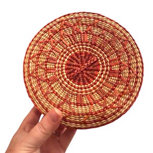 Può includere: Un cestino rotondo intrecciato con un design circolare. Il cestino presenta un colore beige naturale con accenti rosso-arancio. Il design include una spirale centrale e linee radiali, creando una superficie strutturata. Il cestino è tenuto da una mano su sfondo bianco.