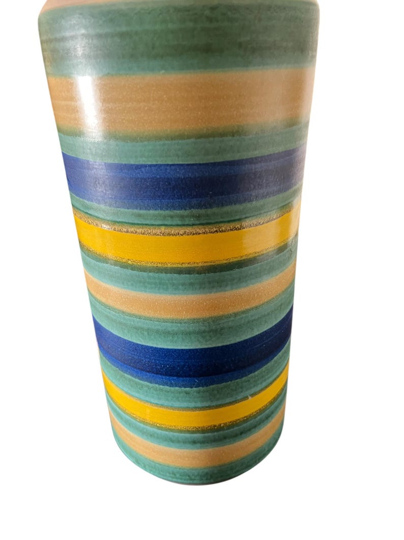 May include: A tall, cylindrical vase with horizontal stripes in shades of green, tan, blue, and yellow. The ceramic vase has a glossy finish and a slightly flared top. The design is a modern take on a classic striped pattern.