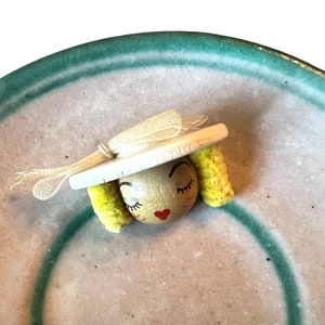 Puede incluir: Una muñeca de madera con un sombrero blanco y pelo de lana amarillo. La muñeca tiene una cara pintada con los ojos cerrados y una boca roja en forma de corazón.