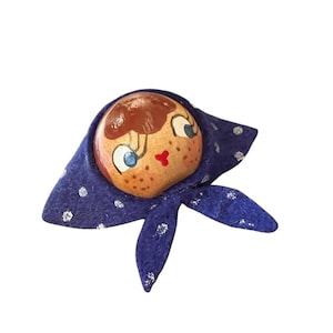 Puede incluir: Una cabeza de muñeca de madera pintada a mano con un pañuelo azul. La muñeca tiene cabello castaño, ojos azules y boca roja. El pañuelo azul está decorado con lunares plateados.