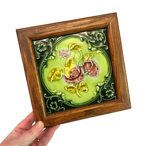 May include: A framed ceramic tile featuring a floral design. The tile has a light green background with a raised floral pattern in yellow and maroon. The frame is made of wood with a natural finish.