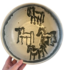 May include: A large, white ceramic bowl with a dark green rim and abstract black and green designs on the inside. The designs resemble stylized animals.