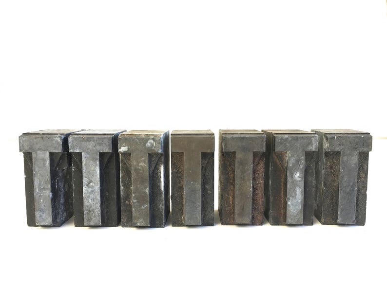 Pu&ograve; includere: Un set di sei blocchi di stampa tipografica in metallo vintage, ognuno con la lettera "T" in uno stile leggermente diverso. I blocchi sono realizzati in metallo grigio scuro e hanno un aspetto usurato e invecchiato.